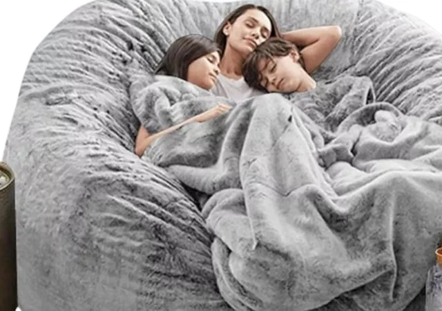 Soft and oversized grey bean bag bed in a cozy bedroom corner, designed for lounging, reading, or relaxing, with plush faux fur fabric and modern style.